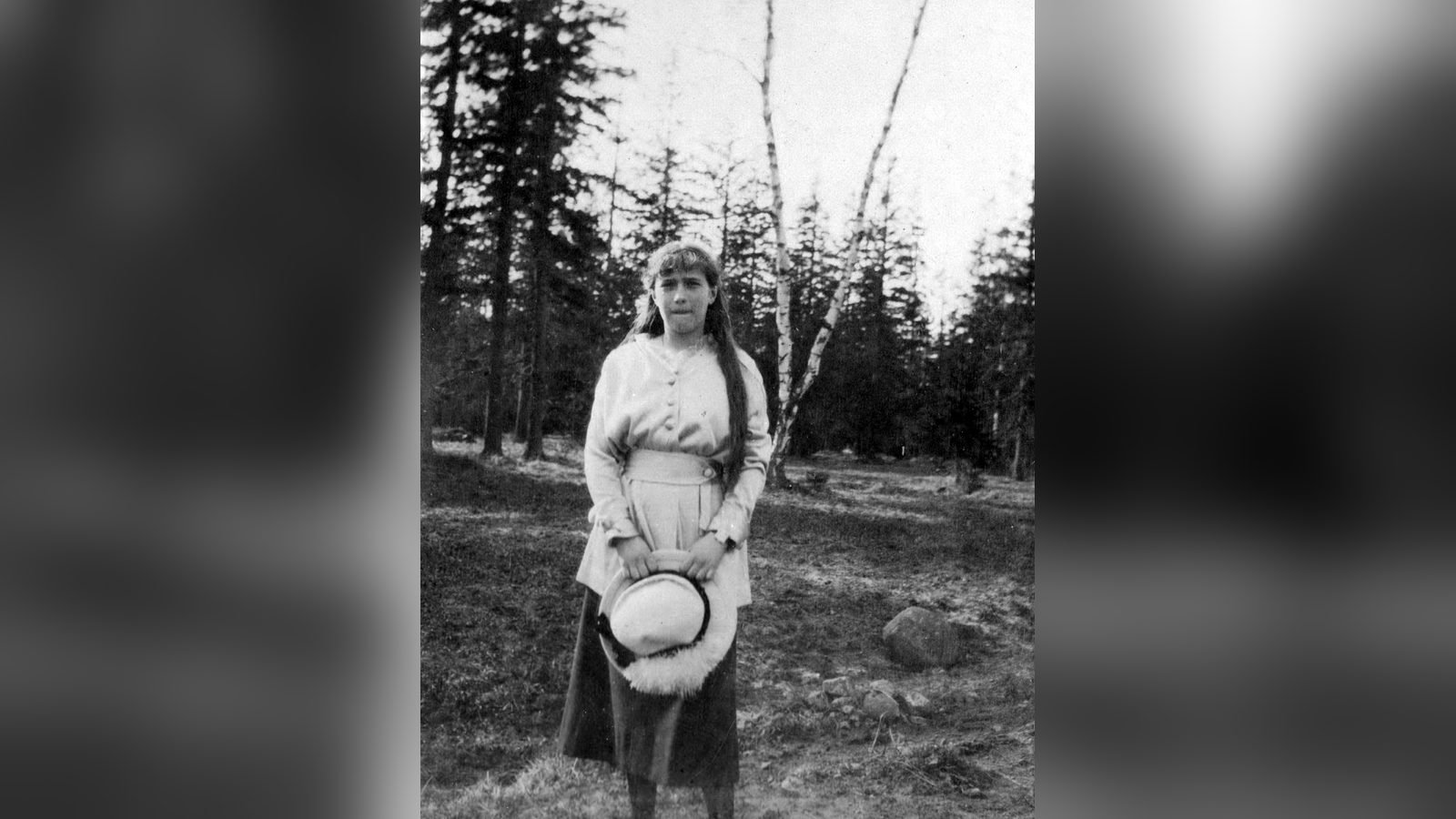La gran duquesa Anastasia Nikoláyevna, de pie en un parque arbolado con un sombrero en las manos, en una fotografía en blanco y negro de 1916