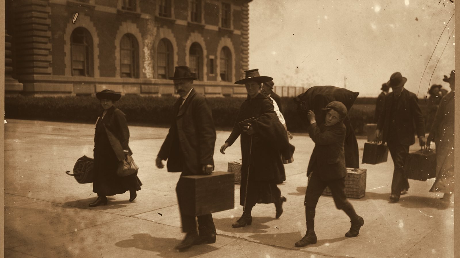 Inmigrantes con maletas y fardos caminando frente al edificio de Ellis Island a comienzos del siglo XX, en tono sepia