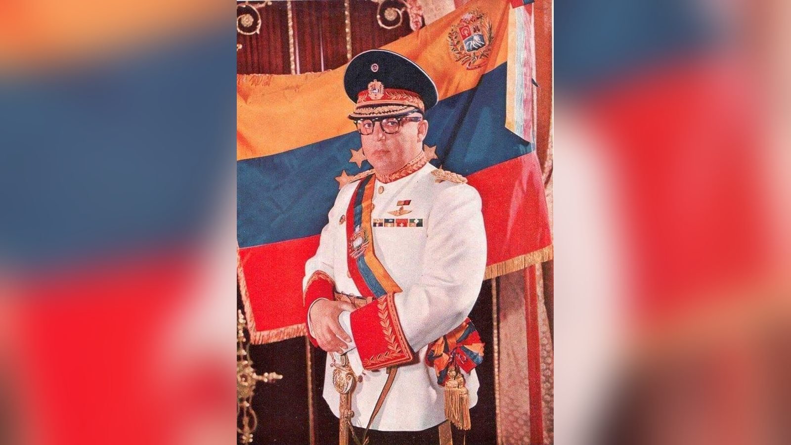 Marcos Pérez Jiménez con uniforme de gala blanco, gorra y banda presidencial, delante de una bandera de Venezuela