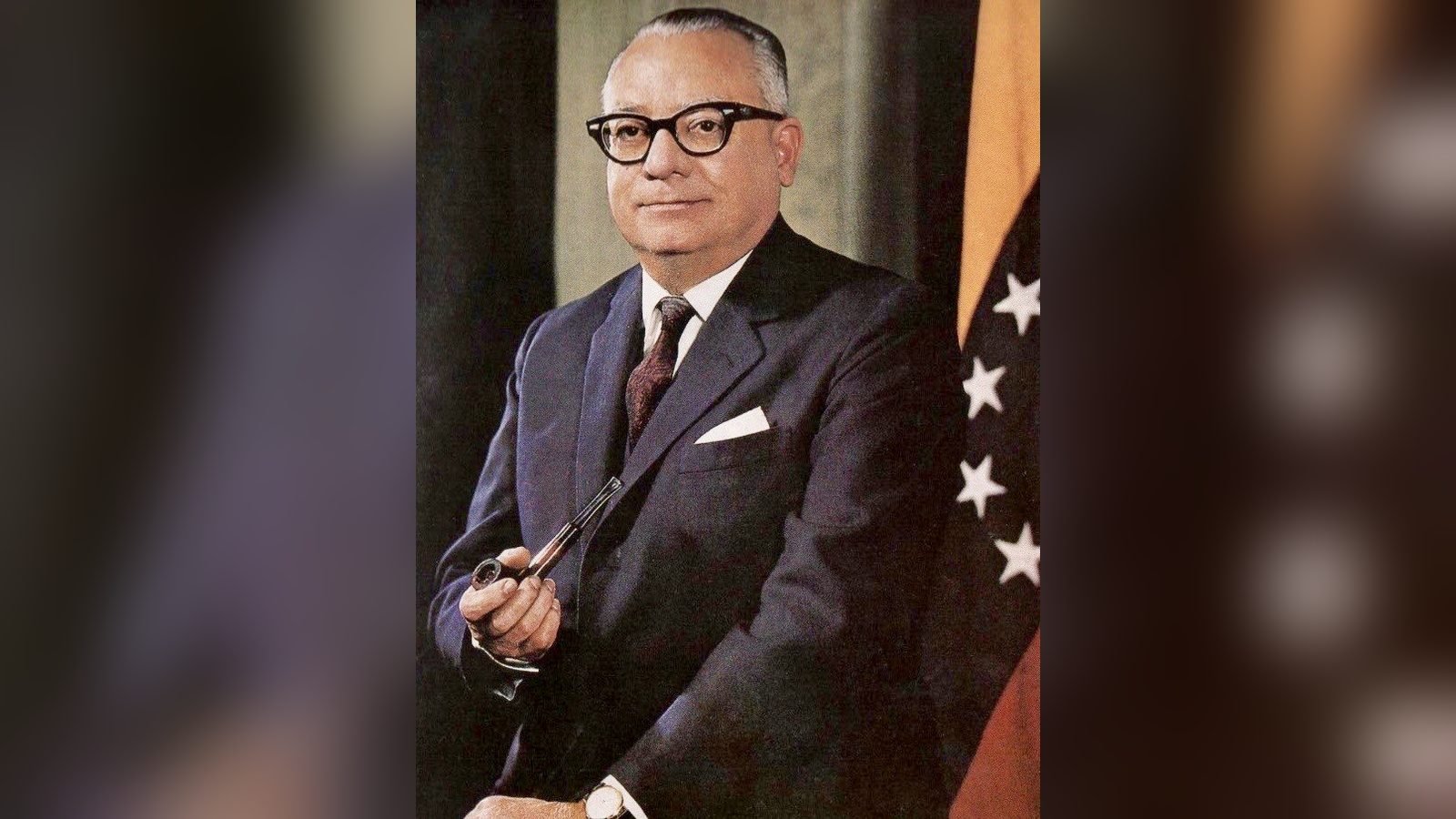 Rómulo Betancourt con lentes y traje oscuro, sentado, con la bandera de Venezuela detrás
