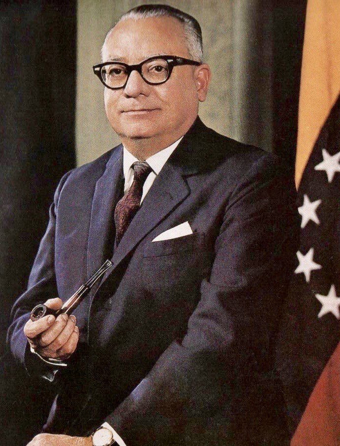 Retrato oficial en color de Rómulo Betancourt con traje oscuro, lentes y una pipa en la mano, junto a la bandera de Venezuela, 1959