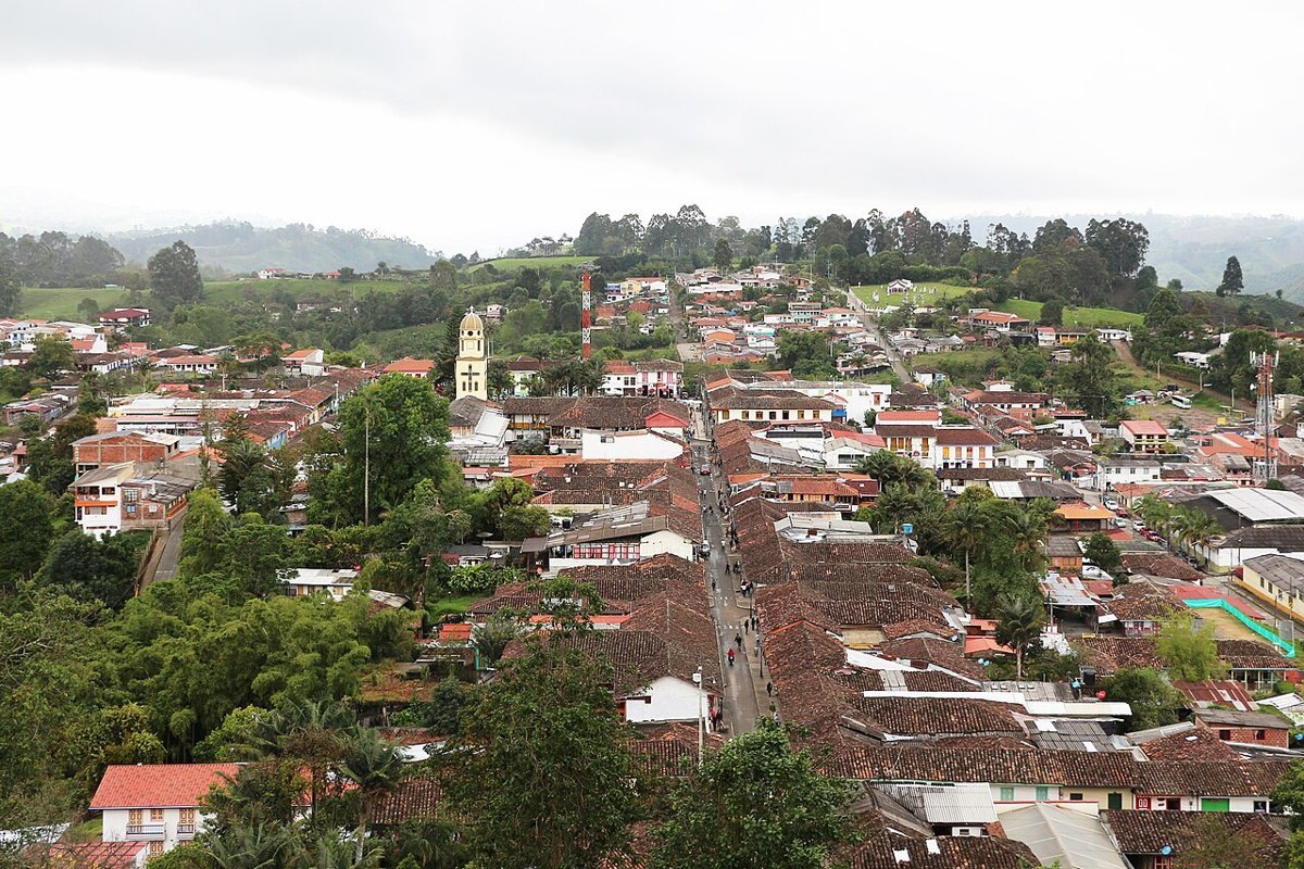 Vista elevada de Salento (Quindío): una calle larga y recta entre casas de tejados de barro, con la torre amarilla de la iglesia a la izquierda y colinas verdes al fondo bajo un cielo nublado