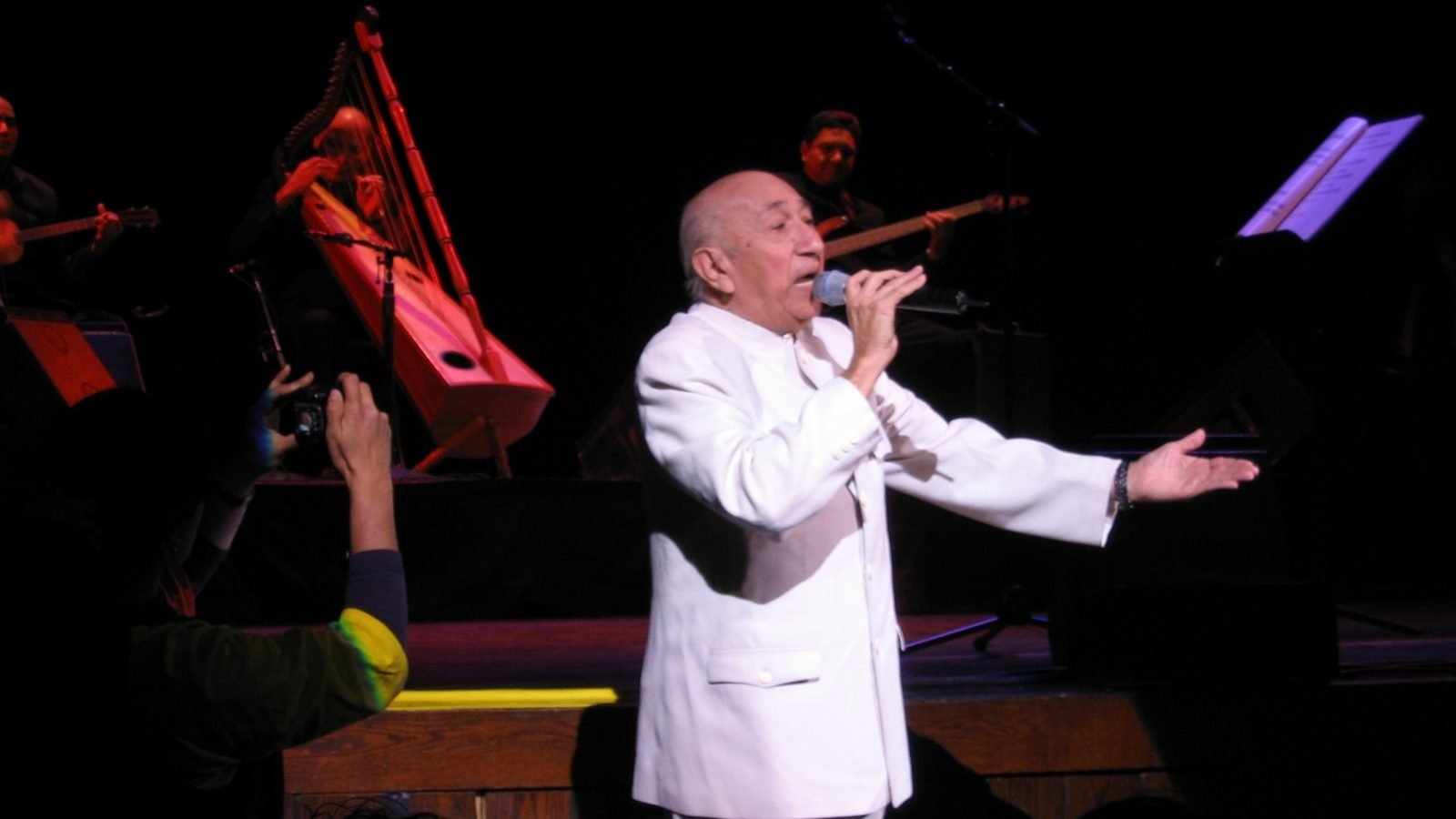 Simón Díaz, vestido de blanco, canta con un micrófono en un escenario con músicos y un arpa detrás